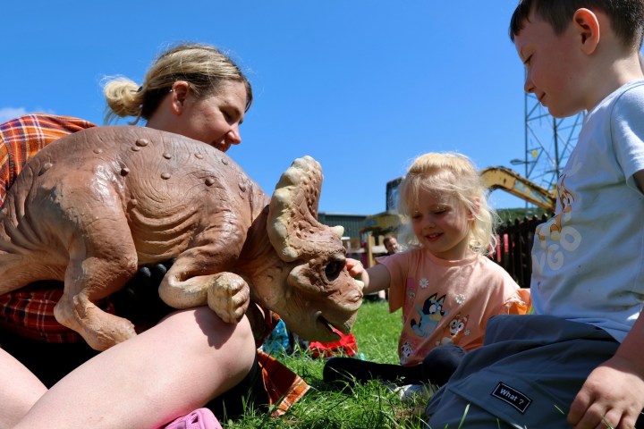 Children and adult interact with dinosaur puppet on grass.