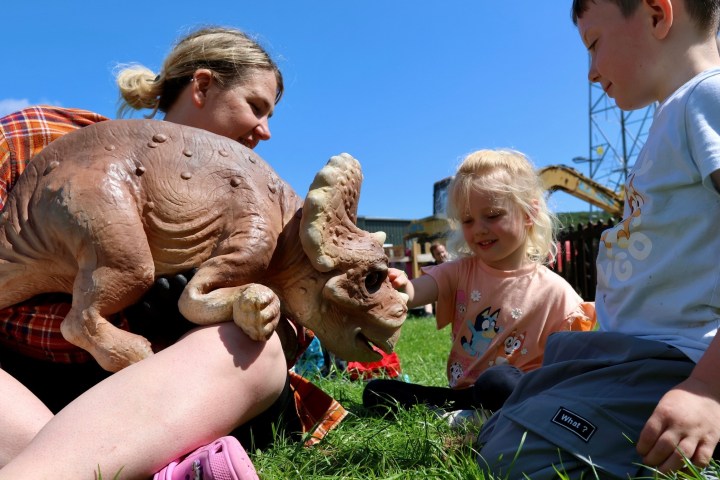 Children play with a dinosaur toy on the grass under a clear blue sky.